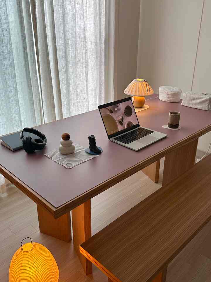 ナチュラルな木目のダイニングテーブルとベンチ、ピンクのレザープレースマットとミニテーブルランプが配置された、雰囲気のあるホームオフィス空間