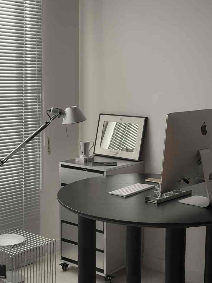 グレーと黒のトーンのホームオフィス、黒い丸テーブルにiMacが置かれ、スタイリッシュなアルミブラインドが配置されたモダンでコンパクトなインテリア