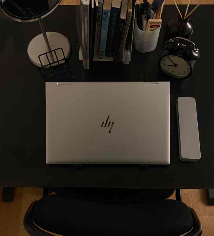 黒いデスクの上にシルバーのノートパソコン、目覚まし時計とブックホルダーが置かれたシンプルなホームオフィス空間