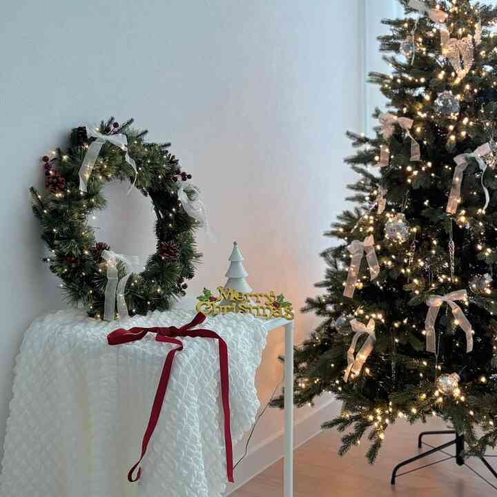 A simple living room with white walls and wood floors featuring a decorated Christmas tree and wreath with ribbon and string lights