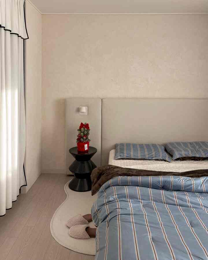 白とベージュのトーンの寝室、ブルーストライプの寝具と黒いスツール上の小さなクリスマスツリーが配置された落ち着いた雰囲気の空間