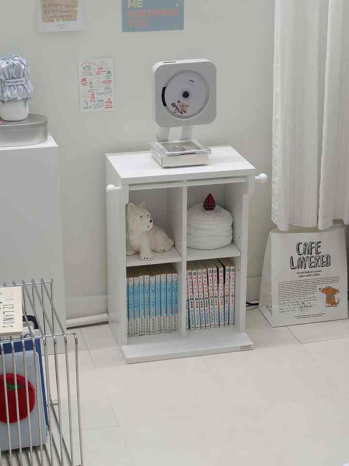 白を基調としたリビングの隅にすっきりとした隙間収納キャビネットが配置され、壁掛け型CDプレーヤーと白いカーテンがある落ち着いた空間