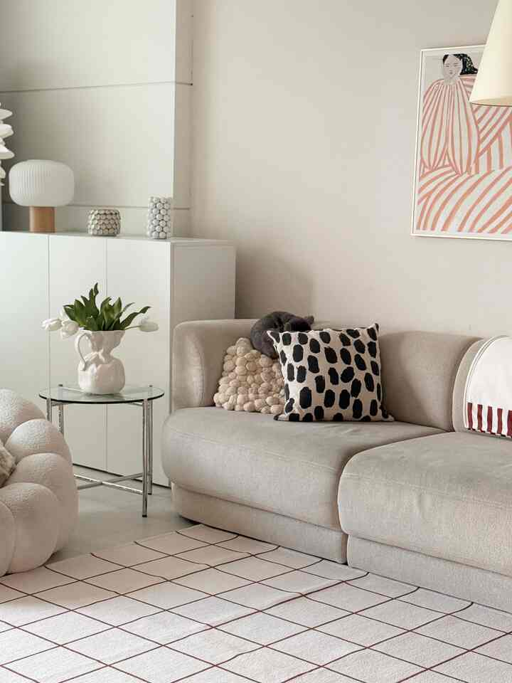 アイボリーとベージュの色調のリビング、快適なソファとクッション、ガラスのサイドテーブルに花瓶、壁に掛けられた抽象絵画が配置された自然でモダンな印象のインテリア