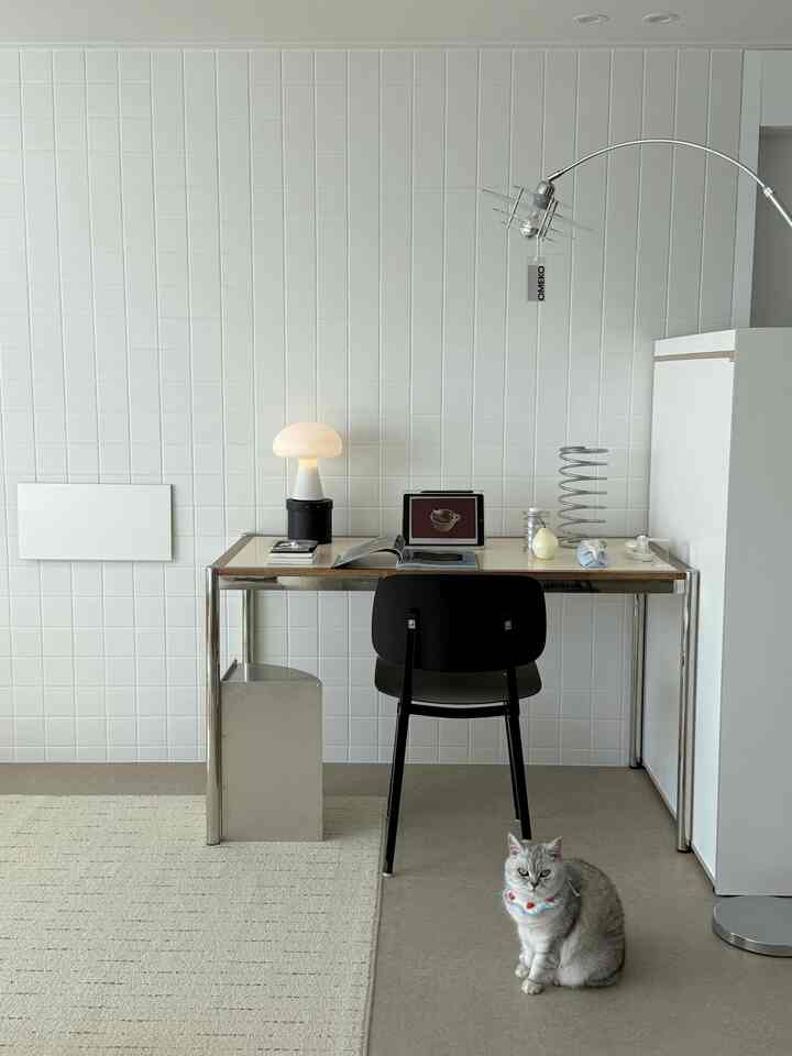 白を基調としたミニマルなホームオフィス、テーブルランプと黒い椅子、前方に座る猫が配置された雰囲気のあるインテリア。