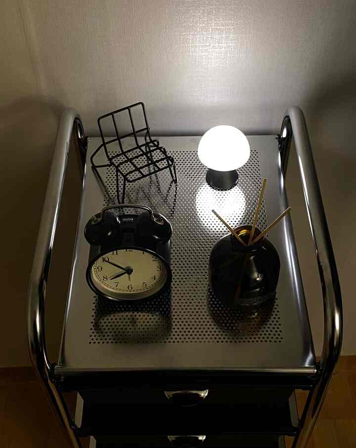 シルバークロームの移動式引き出しトロリーの上に黒い目覚まし時計、LEDキノコランプ、ディフューザーが配置されたモダンでミッドセンチュリーモダンな雰囲気の寝室インテリア