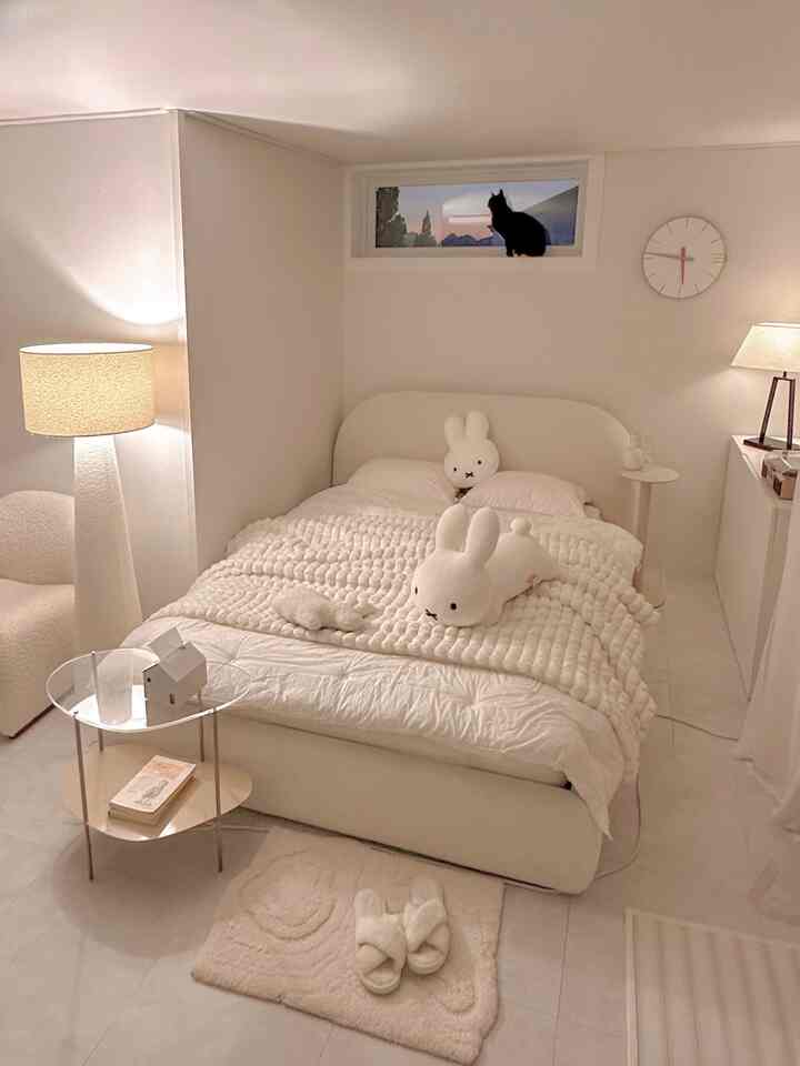 明るい白を基調とした寝室、ベッド、ラグ、ナイトテーブルと照明が配置され、小さな窓に猫が座っている雰囲気のあるインテリア