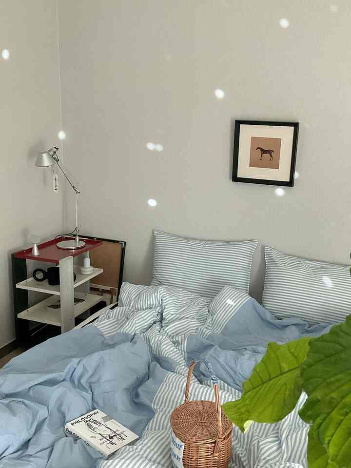 青と白のストライプの寝具があるシンプルでナチュラルな雰囲気の寝室、左側にテーブルランプと額縁が配置されている