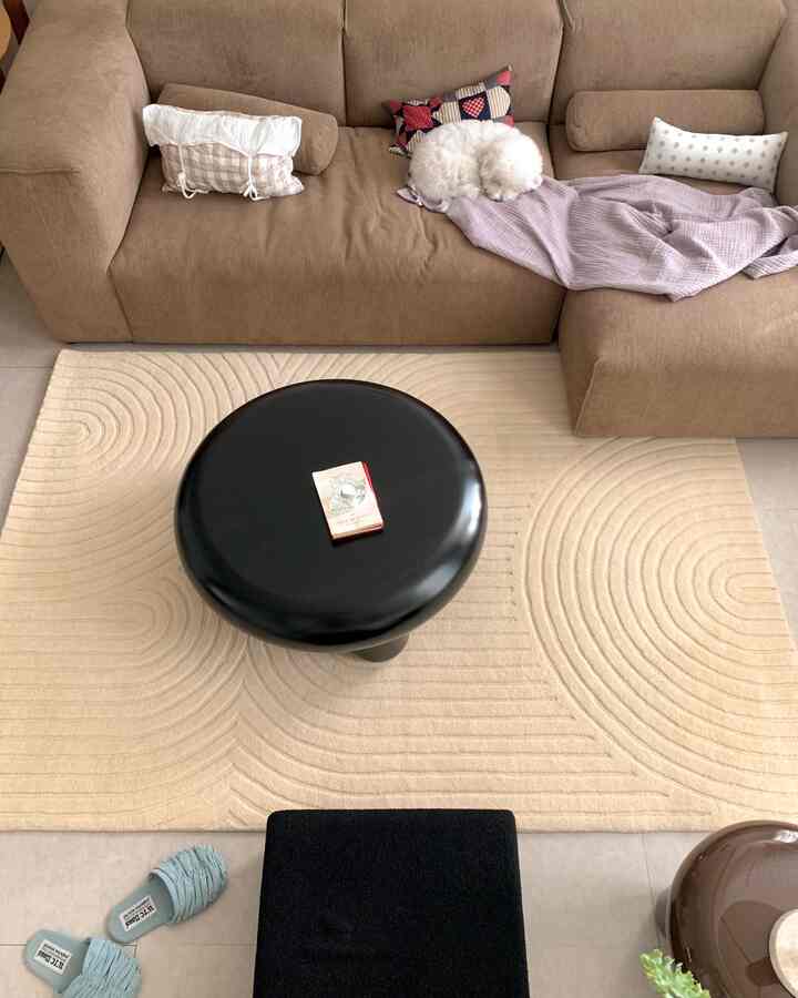 ベージュとブラウンの色調のリビング、大型のL字型ソファと中央に黒い丸テーブルが配置された落ち着いた空間