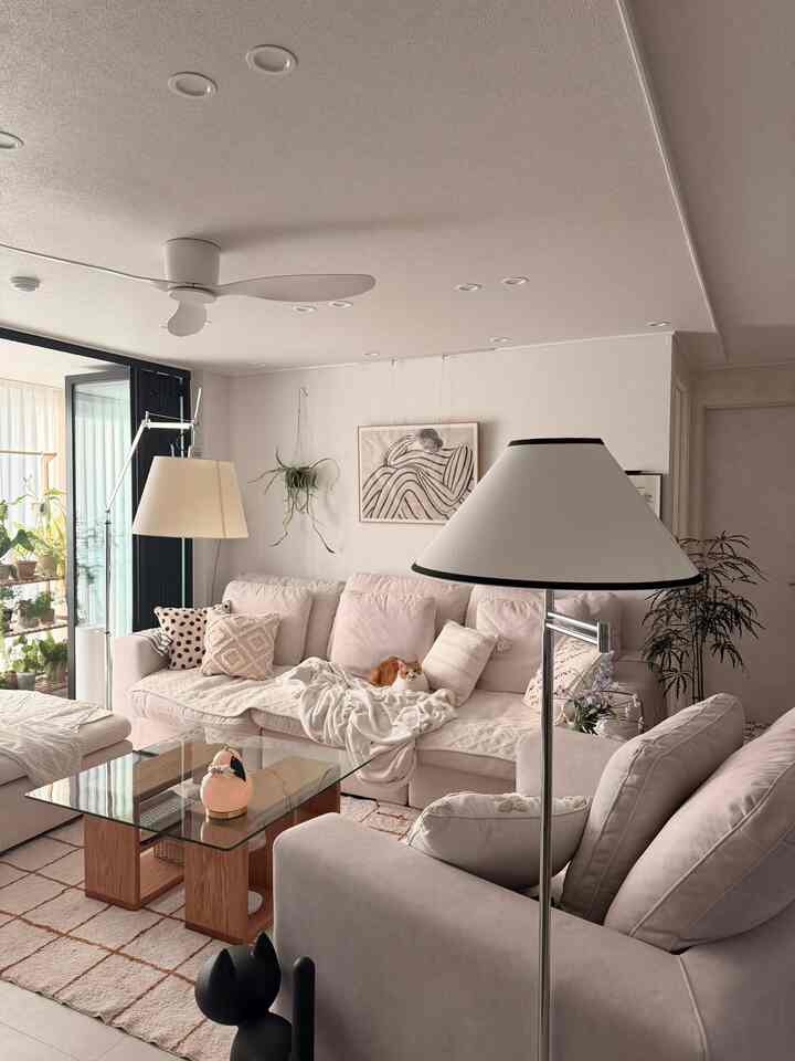 明るいベージュとアイボリーのナチュラルモダンなリビング、広いコーナーソファとガラストップのコーヒーテーブル、猫と多彩なクッションが配置された快適な空間