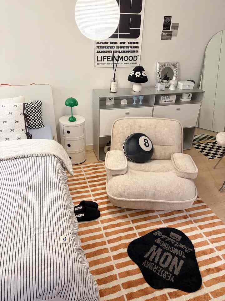 ベージュとホワイトのトーンのワンルーム寝室、ベージュのアームチェアソファ、無彩色のラグ、センスのある照明とポスターが配置された雰囲気のあるインテリア