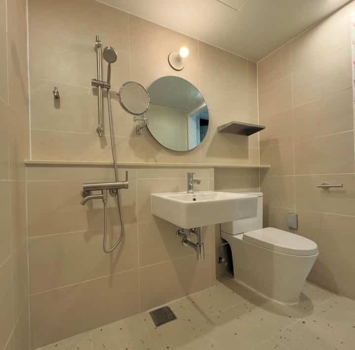 ベージュのタイル壁と床を持つ収納なしのバスルーム、丸い鏡、露出型洗面台、スカート型トイレが配置された清潔感のある空間