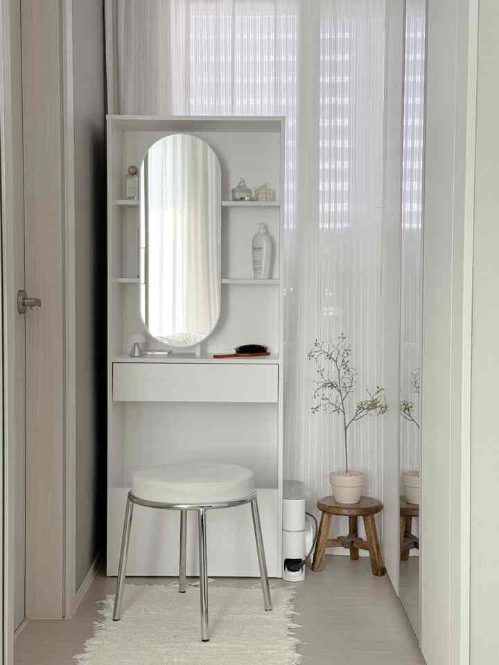 ホワイトトーンのコンパクトな寝室、縦長の化粧台とスツール、木製のスツールと植物が配された清潔で居心地の良いインテリア