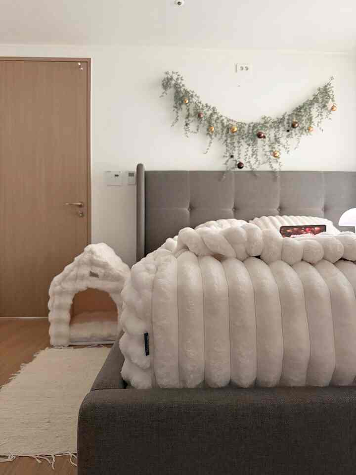 白とグレーのトーンの寝室、大きなベッドとふわふわのブランケット、左側にペットハウスが置かれた居心地の良いインテリア