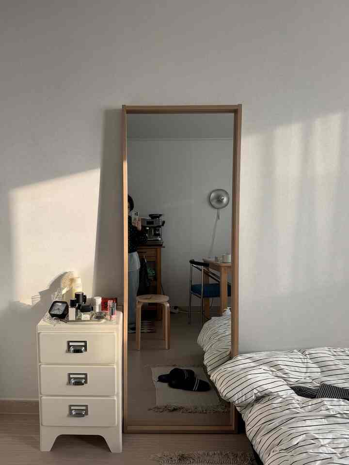ホワイトの壁と木目調の鏡と家具のある寝室、ベッドカバーとスツールが配置されたシンプルな空間