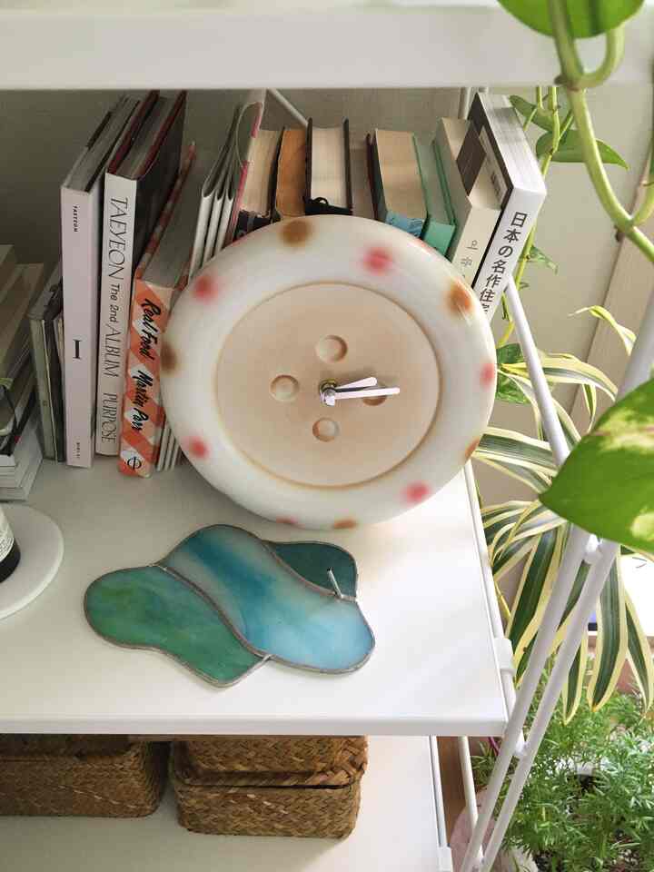 ベージュと白の色調のワンルームの棚に、ボタン型の時計や植物、本が配置された雰囲気のあるインテリア