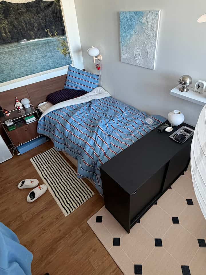 Compact bedroom with white walls and wood-tone flooring, featuring blue striped bedding, nightstand, and wall sconce creating a cozy atmosphere