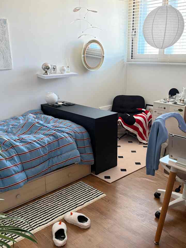 Modern bedroom with white walls and window, featuring a black storage cabinet centrally placed, a bed with blue striped bedding, small rugs, desk, and chair creating a harmonious atmosphere