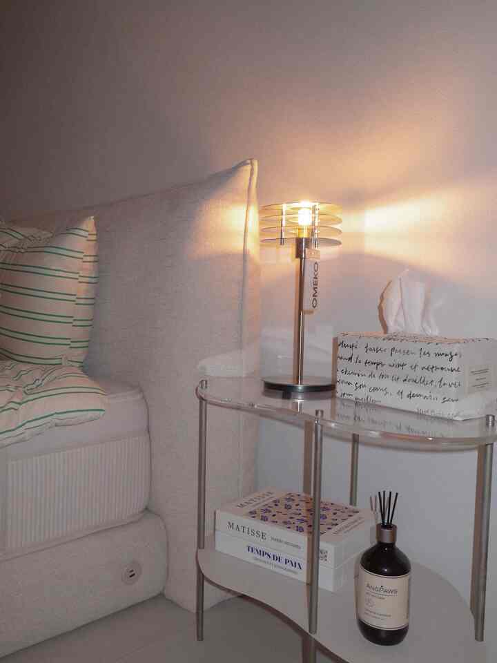 アイボリーと白の色調の寝室、アクリル製の棚とテーブルランプ、ディフューザーが配置された居心地の良い空間