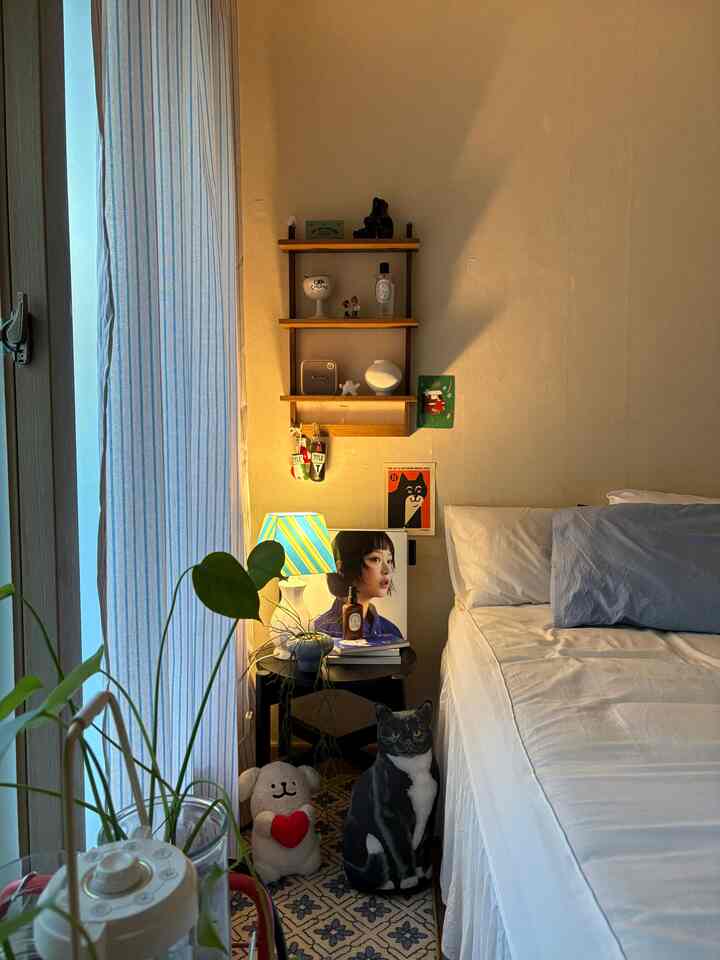 柔らかなアイボリー色の壁とブルーストライプのカーテンが調和する一人暮らしの寝室、木製の壁棚とかわいいペットモチーフの小物が配置された雰囲気のあるインテリア