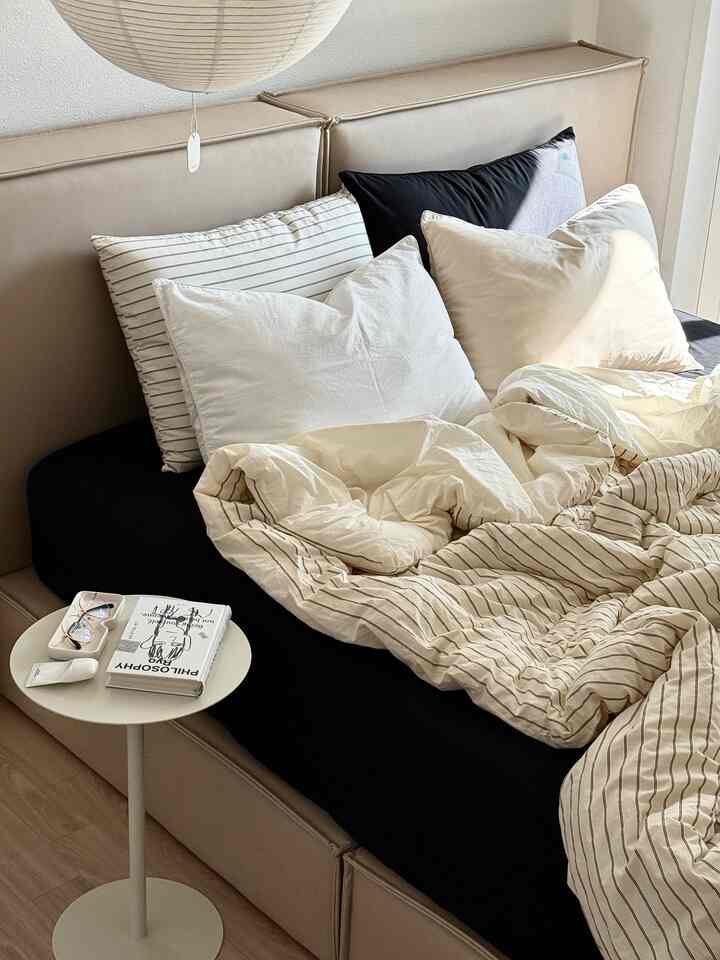 ベージュトーンの寝室、中央にベッドと枕があり、左側に丸いナイトテーブルが配置されたシンプルモダンな空間