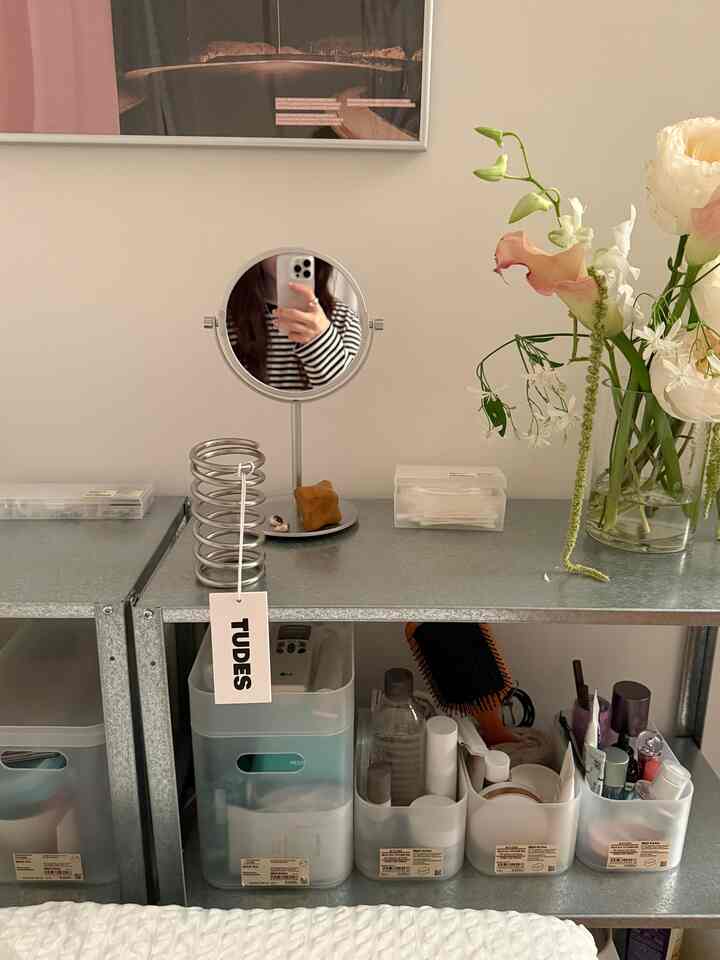 Simple and clean space featuring a silver galvanized metal shelf with a round mirror, transparent plastic storage boxes, and a vase with flowers