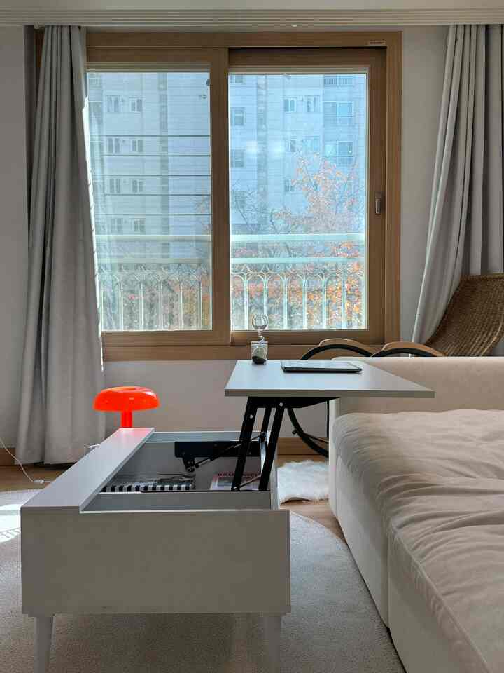 明るい白と茶色のトーンのリビング、窓際にソファとリフトアップ式コーヒーテーブルが配置され、シンプルで実用的な雰囲気のあるインテリア