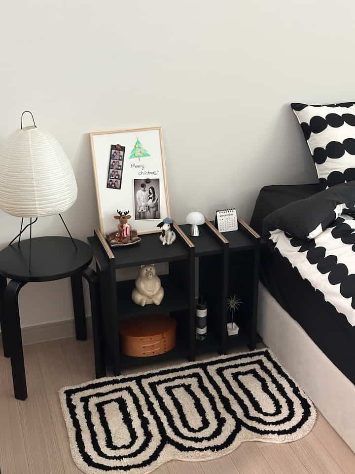 白い壁と黒い家具が配置されたシンプルな寝室、パターンのラグと小さなテーブルランプが印象的な空間