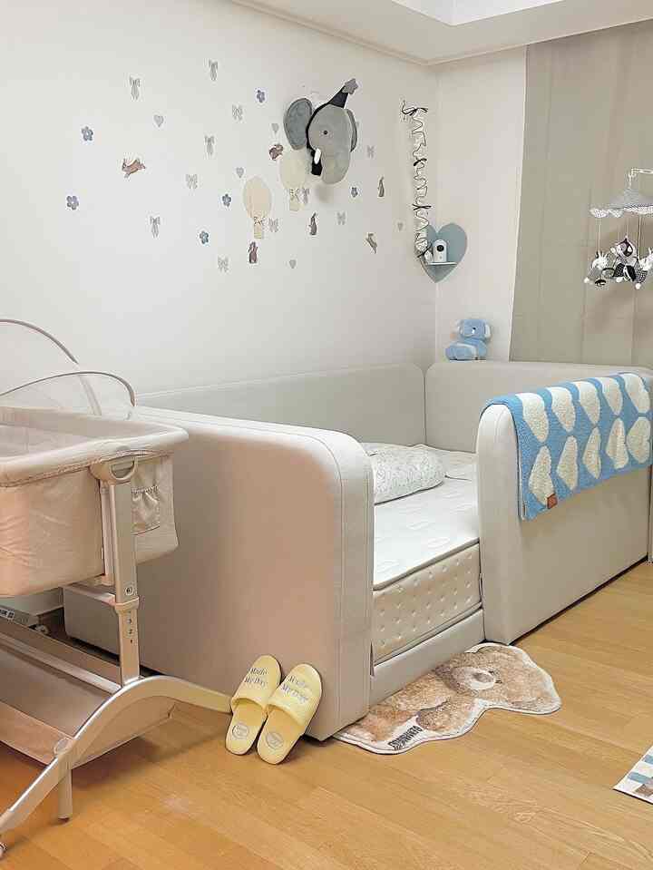 Beige-toned baby room featuring a sofa bed, baby crib, wall decorations, and a teddy bear rug creating a soft and cozy atmosphere