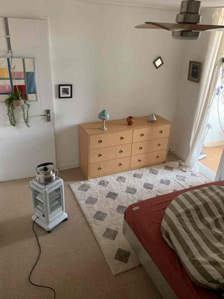 ベージュとホワイトの色調の寝室、木製キャビネットとヒーター、ストライプの寝具が配置されたヴィンテージ感のあるアパートインテリア