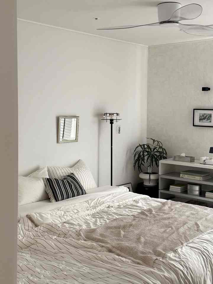 白とベージュのトーンが広がる寝室、中央に大きなベッドと枕、隣に棚とヴィンテージ風フロアランプが配置された雰囲気のあるインテリア