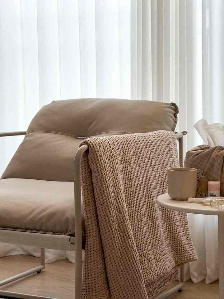 ベージュとブラウンの色調のリビング、快適なアームチェアとラウンドサイドテーブル、白いカーテンが配置された雰囲気のあるインテリア