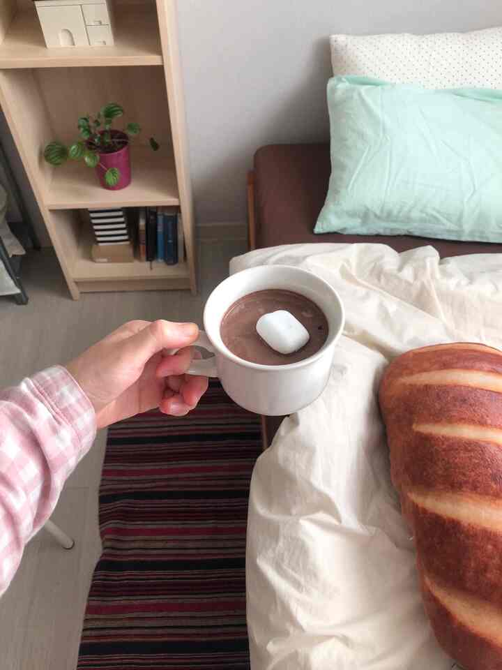 A cozy bedroom with warm white and brown tones featuring a wooden bookshelf and a hand holding a mug, creating a relaxing atmosphere