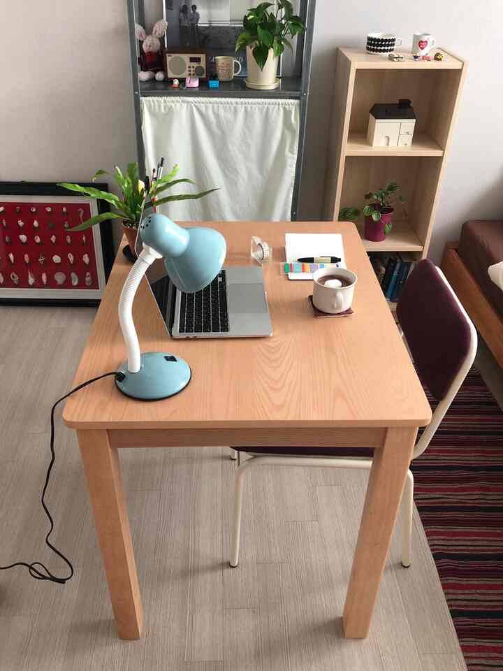 A white-walled home office space with wood-tone furniture, featuring a desk with a sky blue lamp and laptop, creating a cozy work area
