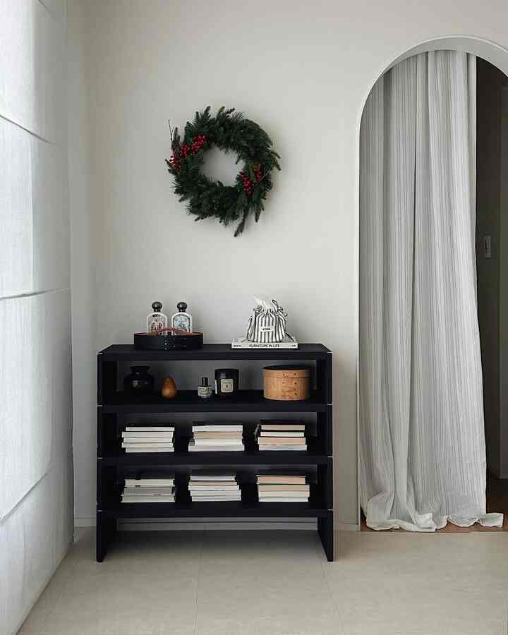 Modern bedroom with white walls and curtains, featuring a black bookshelf and a Christmas wreath arranged neatly