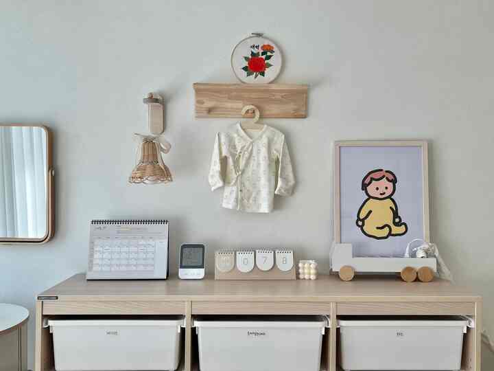 クリーム色と木目調の子供部屋、赤ちゃんの服、カレンダー、木製棚、ラタンの壁照明が配置された雰囲気あるインテリア