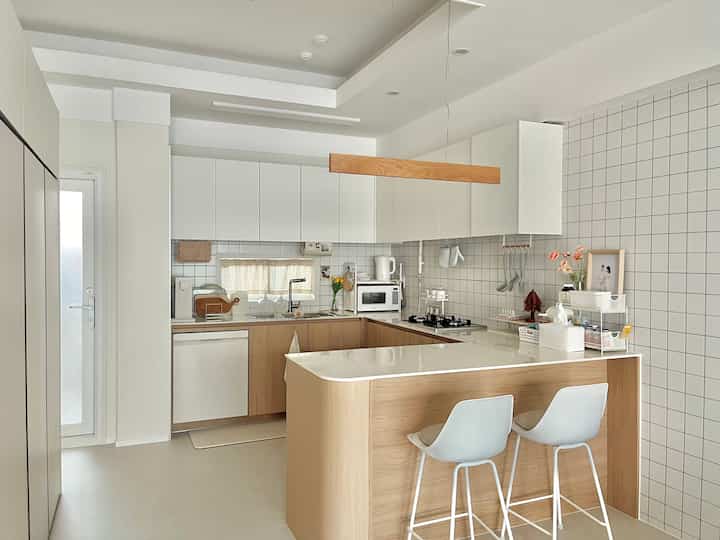 木目調とクリーム色の温かみのあるU字型キッチン、ペンダントライトとダイニングチェアが配置されたモダンで自然な雰囲気の空間