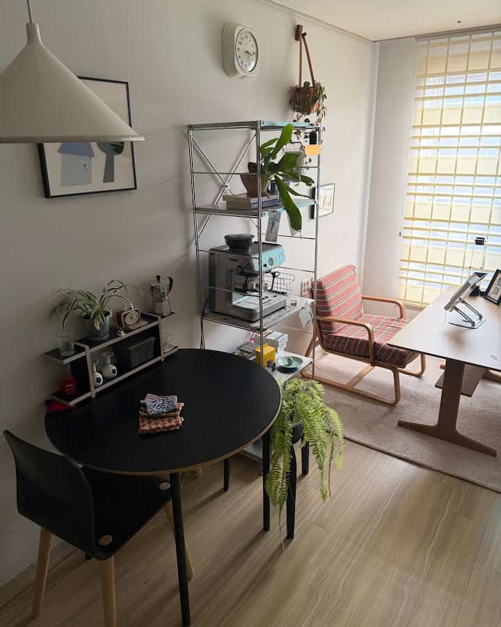 Beige and black toned living and dining space featuring wooden desk, black dining table, plants, and coffee machine in a clean modern interior
