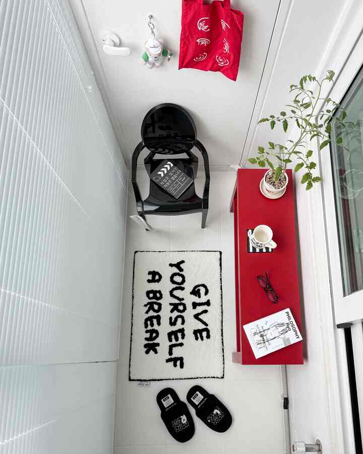 A narrow balcony space in white and red tones featuring a black chair, lettering rug, and a plant with a simple modern vibe