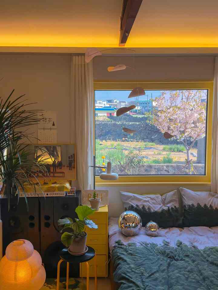 A cozy bedroom with warm yellow lighting, featuring a window and natural plants arranged around the bed