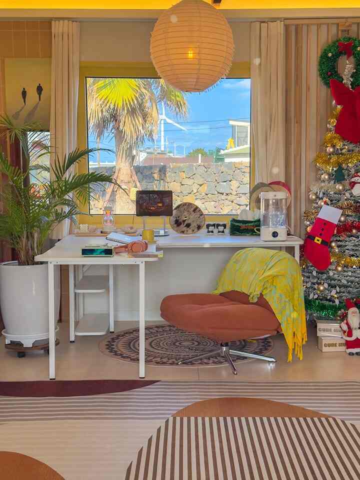 Bright-toned home office space featuring central desk and swivel armchair, Christmas tree with pet stocking on right creating a cozy living room atmosphere