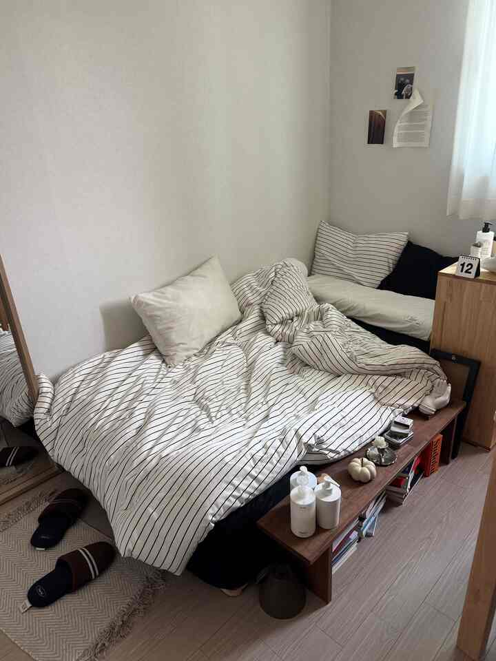 ホワイトと木目調の縦長寝室、ストライプの寝具と木製の棚が配置されたシンプルで落ち着いたインテリア