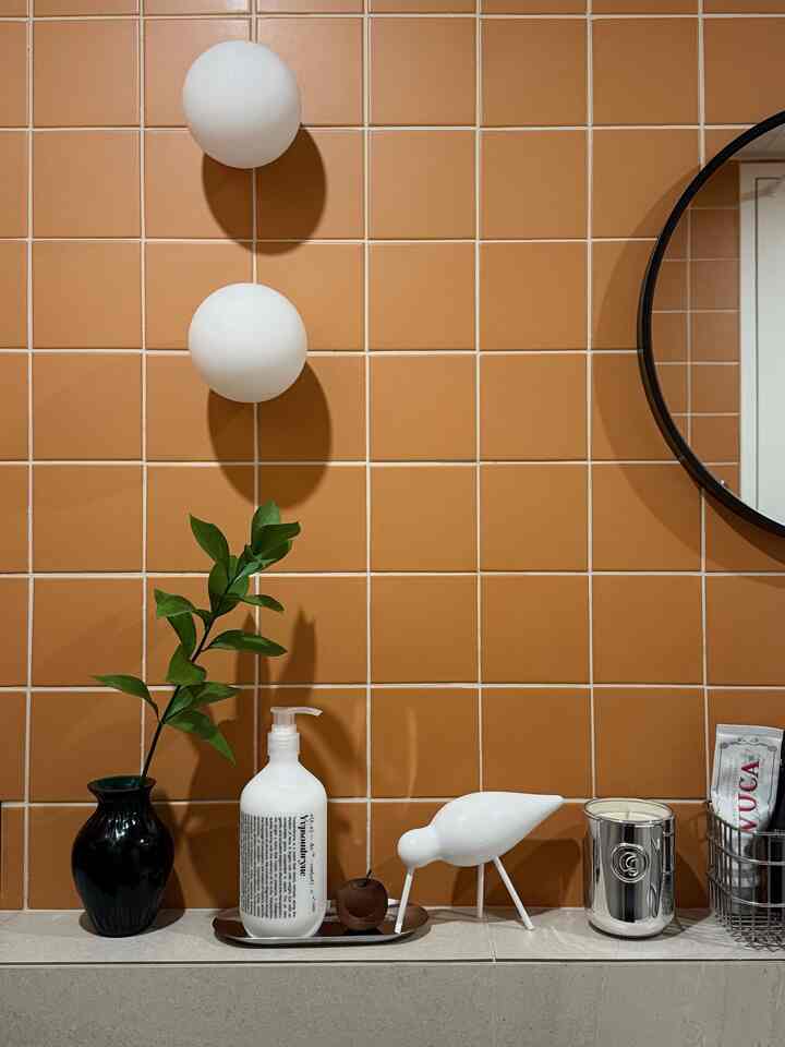 Bathroom space with orange tiled wall, white lights, a round mirror, and decorative items arranged on a shelf