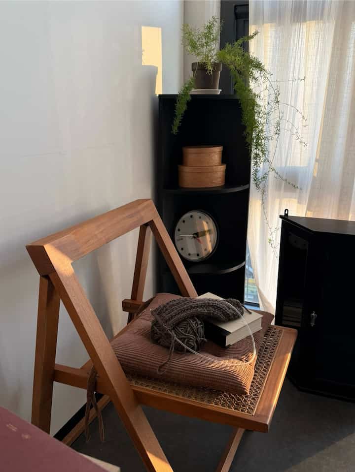 Cozy corner space featuring a wood-toned chair, black shelf with a clock and a plant under soft natural light