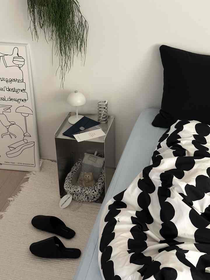 White-walled bedroom featuring natural tone rug, black patterned bedding, and a nightstand with table lamp in a simple modern setting