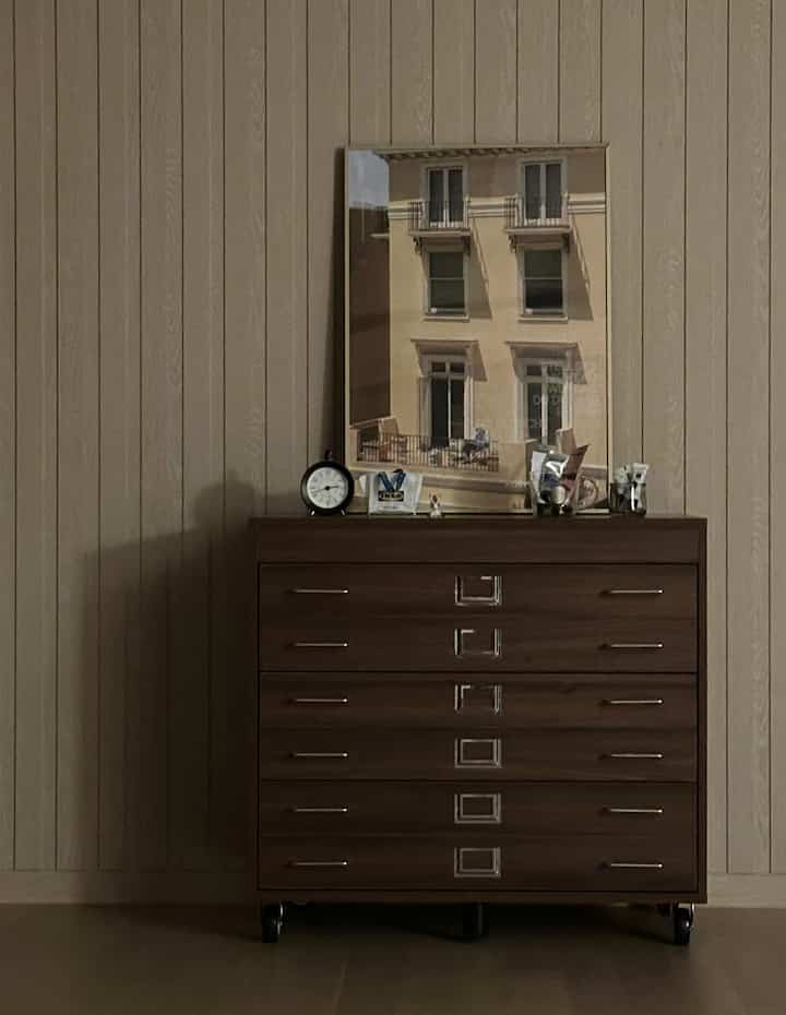 ブラウンとナチュラルカラーの壁を背景に、ドレッサーの上に置時計とポスターが配置された落ち着いた雰囲気のインテリア