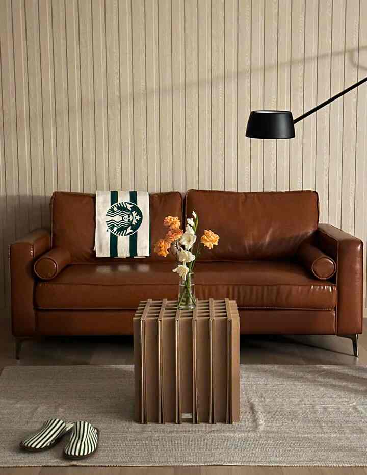 アイボリー色の木目壁とブラウンの革ソファが配置されたナチュラルモダンなリビング、中央にユニークなコーヒーテーブルとフロアランプが置かれている