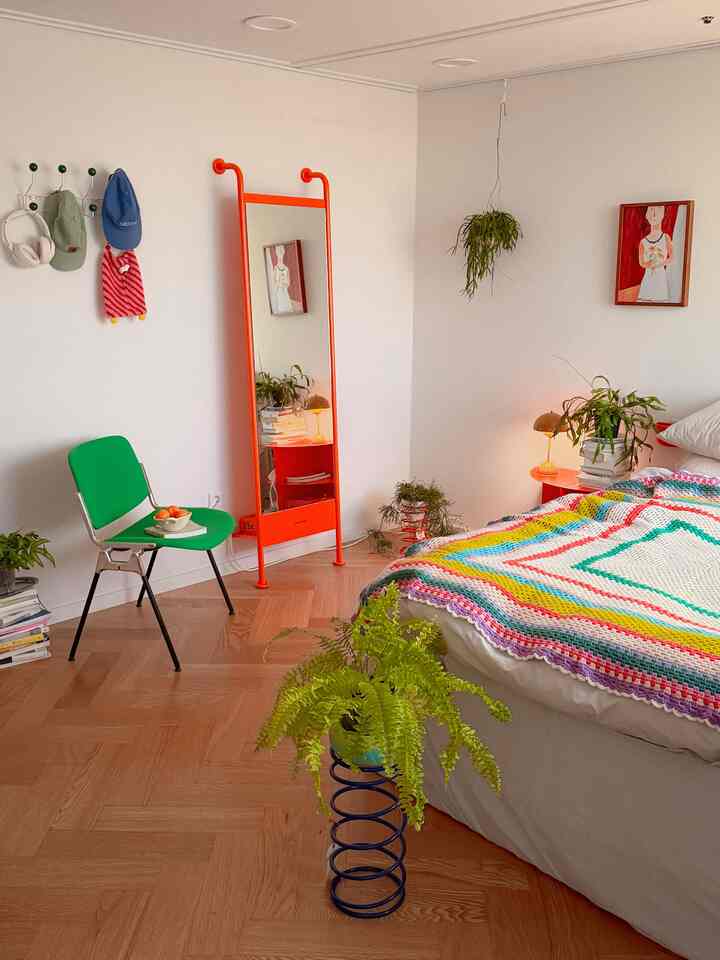 Bright bedroom with white walls and vibrant orange mirror, decorated with lush plants and colorful bedspread
