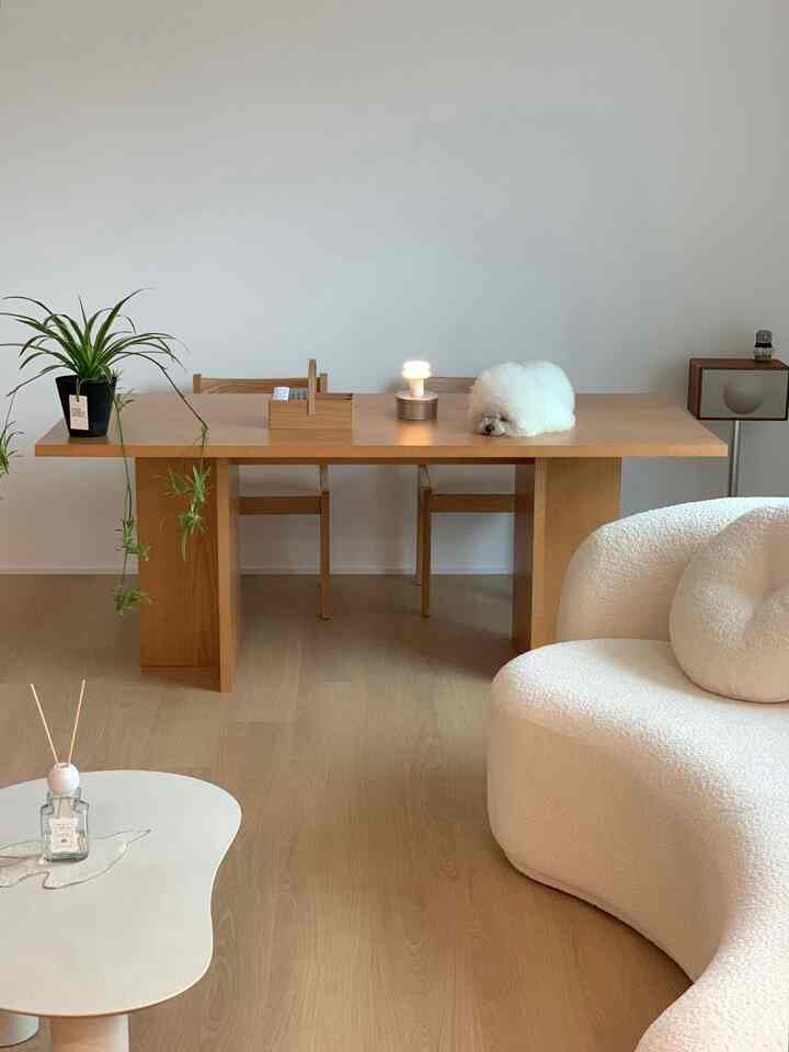 Warm wood-tone living room featuring a large wooden dining table, white curved sofa, and a small white dog resting on the table, creating a cozy atmosphere