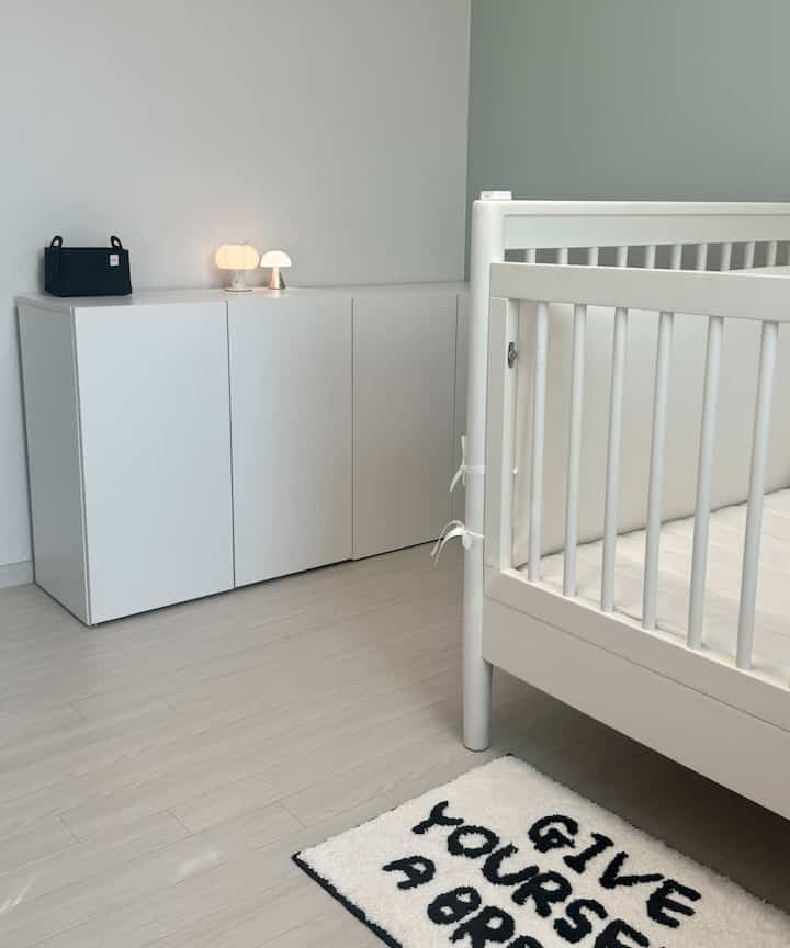 白と薄いグレーの色調の子供部屋、清潔感ある白いベビーベッドと収納キャビネットが配置された印象的な空間