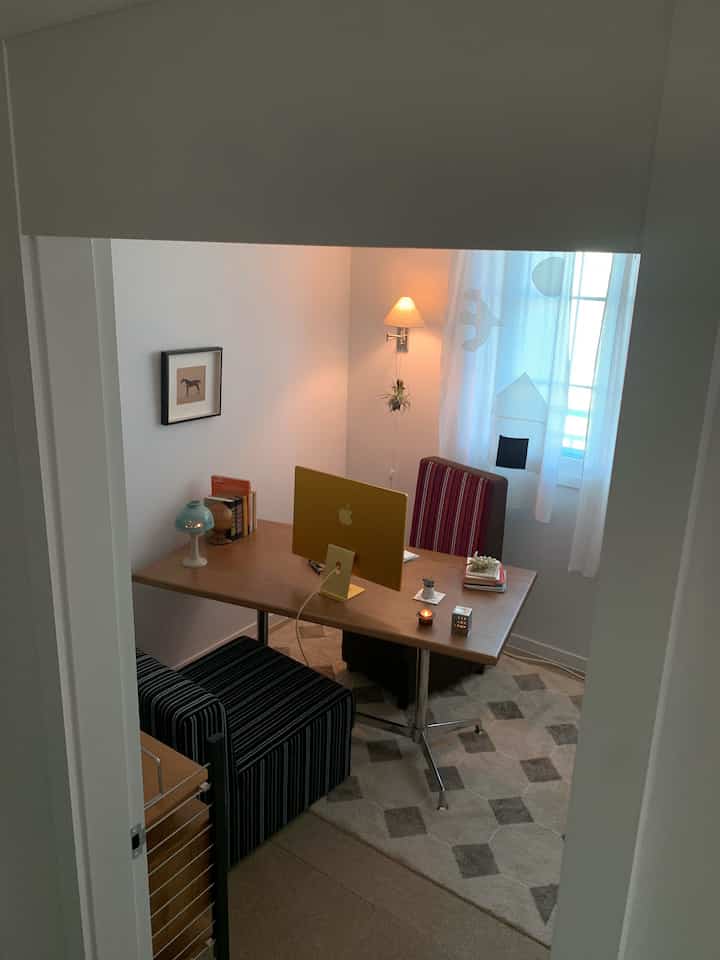 Cozy home office study with white walls and curtains, wood-toned desk featuring a yellow computer
